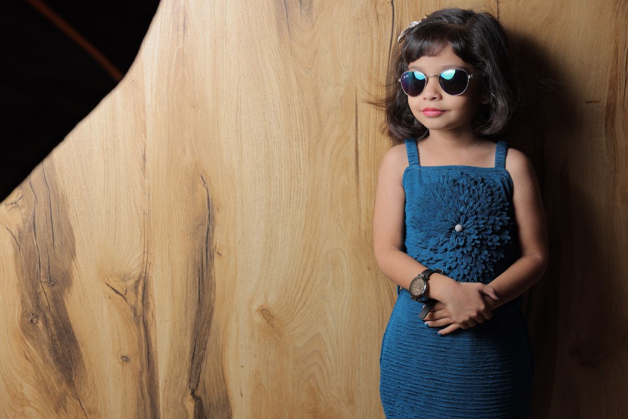 Cute and fashionable young girl wearing sunglasses and a blue dress against a wooden backdrop.