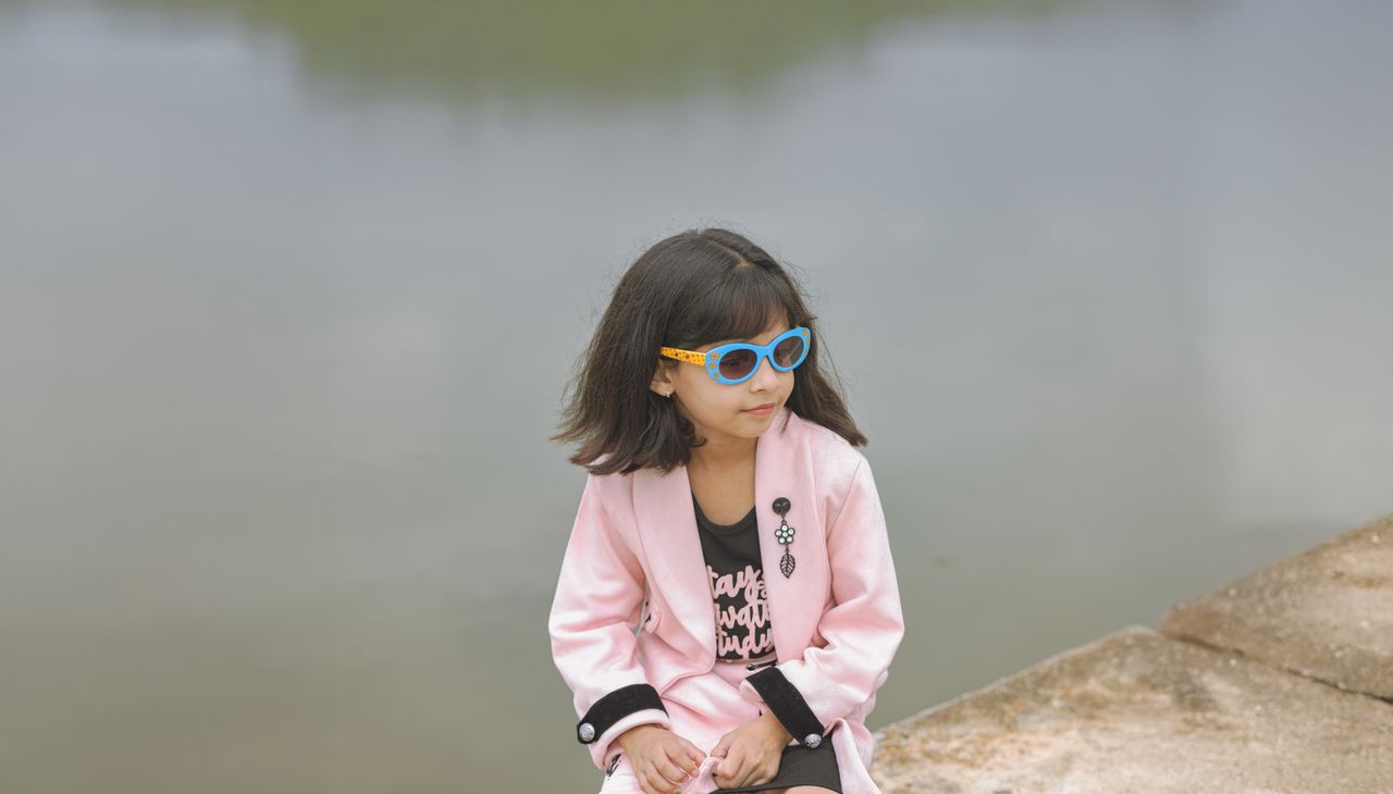 A young girl in sunglasses and a pink jacket sits by a serene lake, embodying youthful fashion.